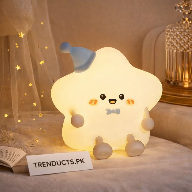 A glowing star-shaped nursery lamp with a cute face wearing a blue hat, sitting on a bed next to a Trenducts.pk card.